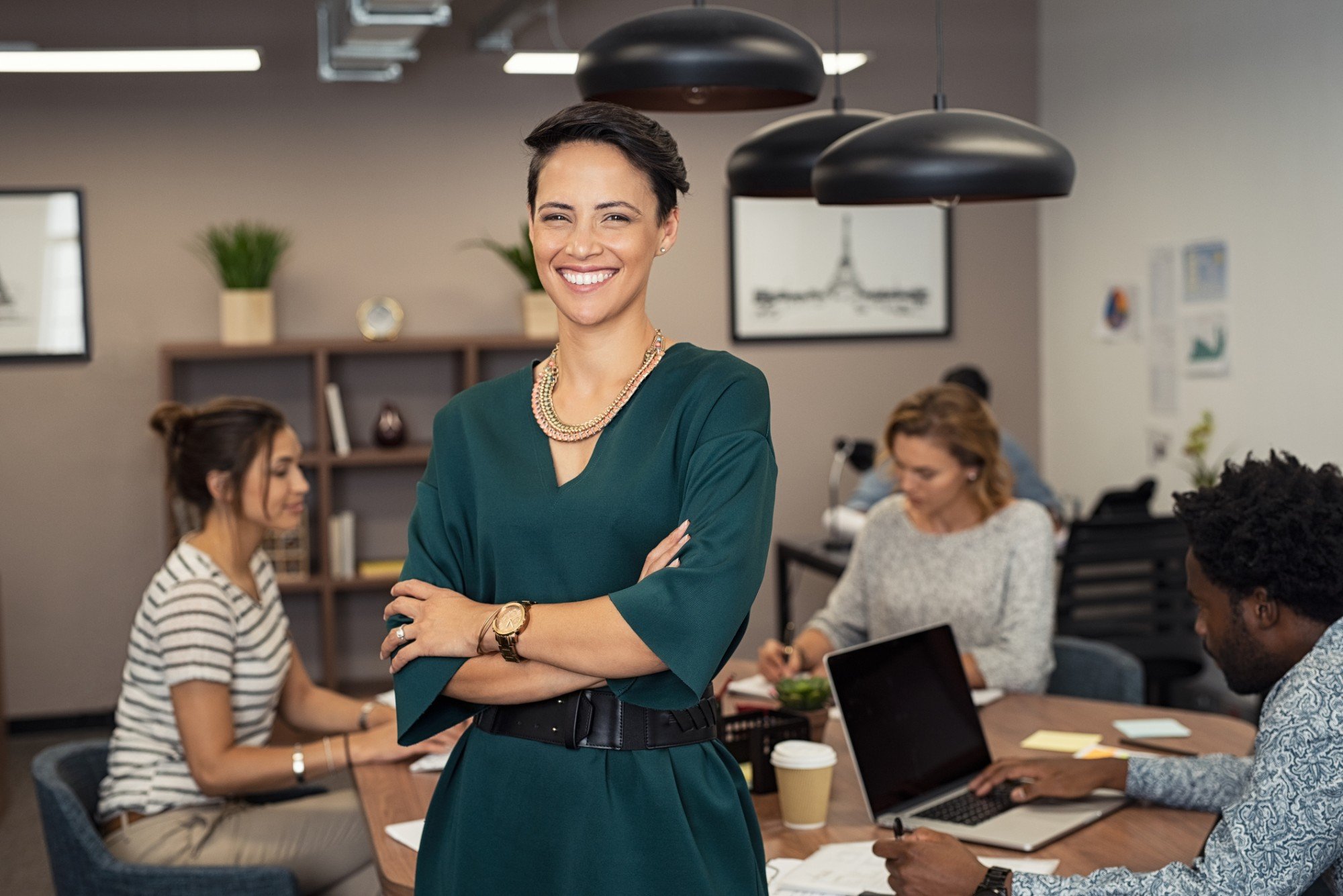 A young black businesswoman smiles and stands confidently with her arms crossed while several other businesspeople work at a roundtable in the background.