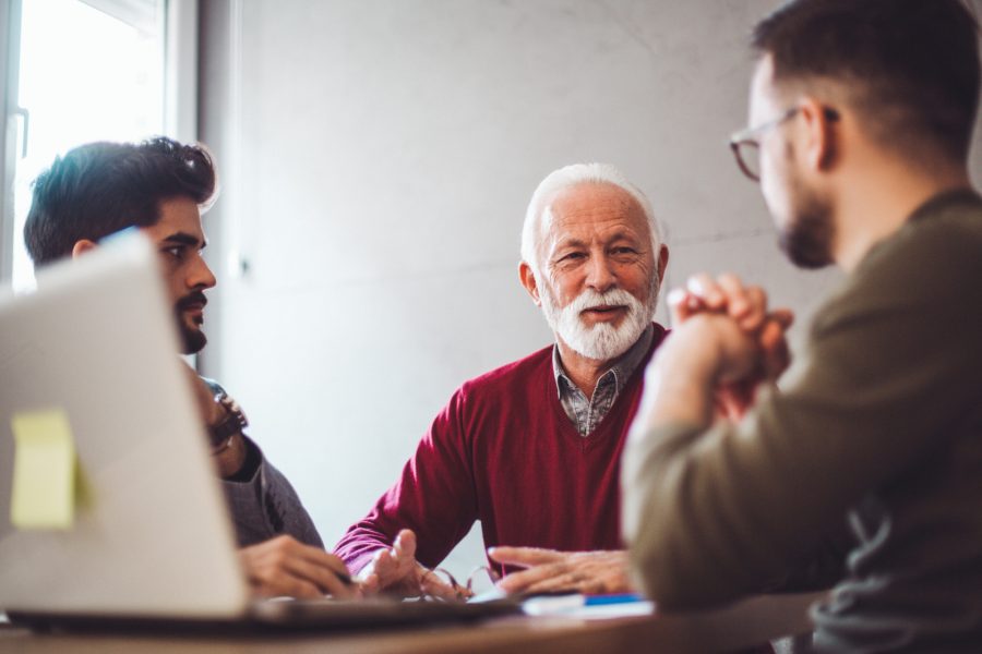 An older caucasian man sits at a desk with two younger men who may be his sons while they have a discussion.