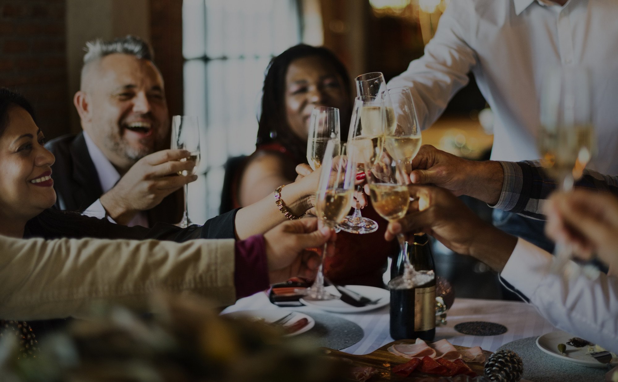 Several business people sit at a table celebrating and doing a cheers with glasses of champagne while laughing happily.