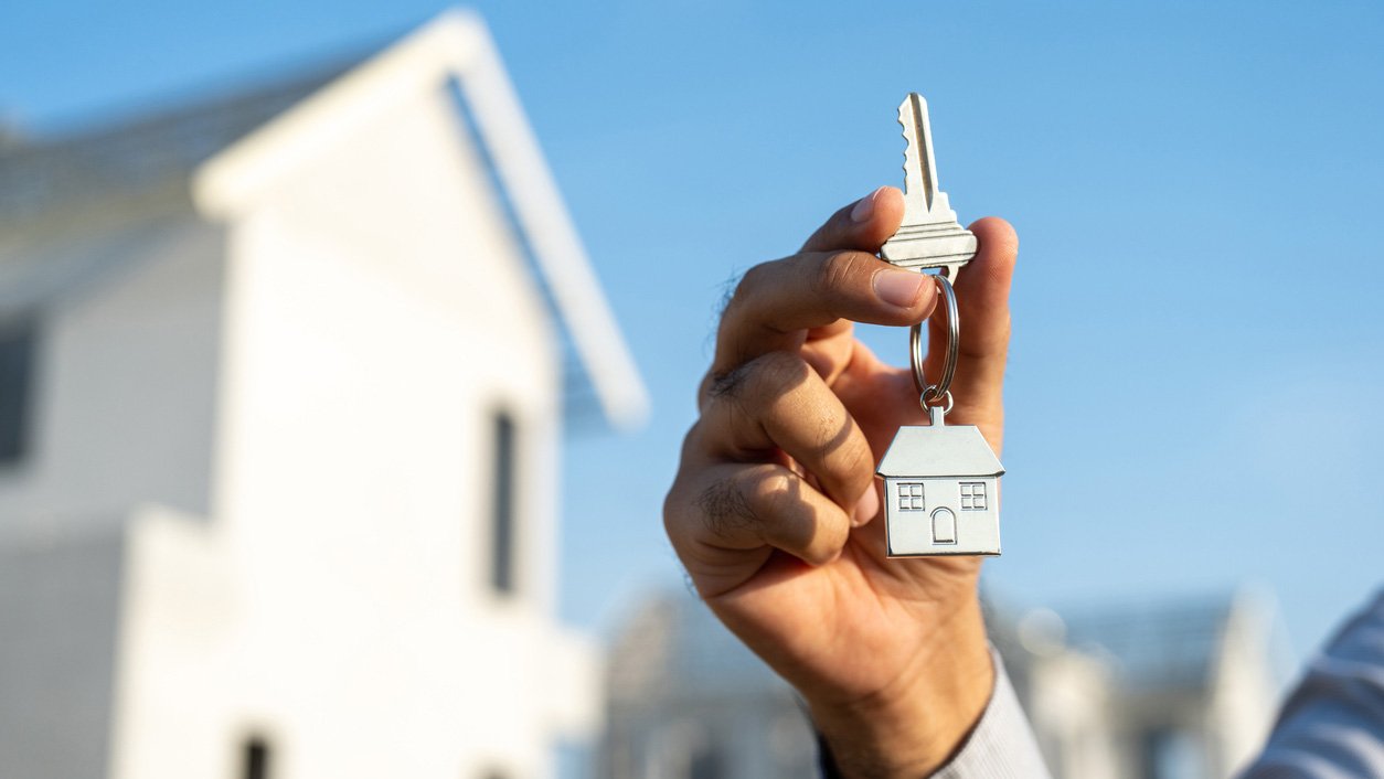 A man's hand holds up the house key to a new laneway house in the background of the photo.