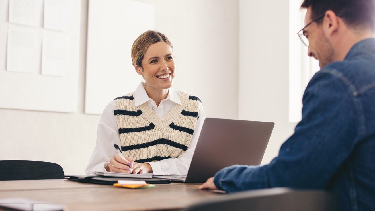 A female business advisor sits at a desk with a laptop taking notes and smiling talking with a client.