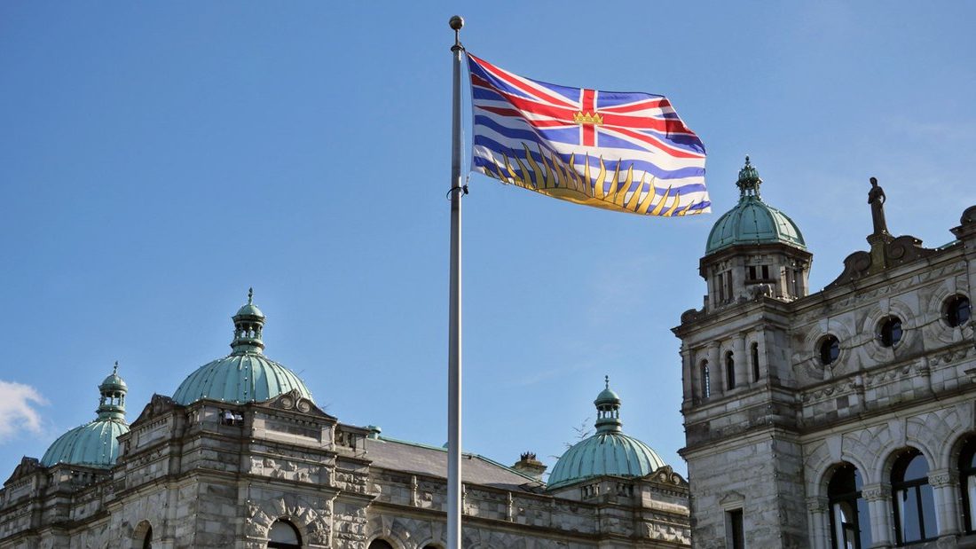 The British Columbia flies on a flagpole, with blue skies and the BC Parliament building in Victoria, BC in the background.