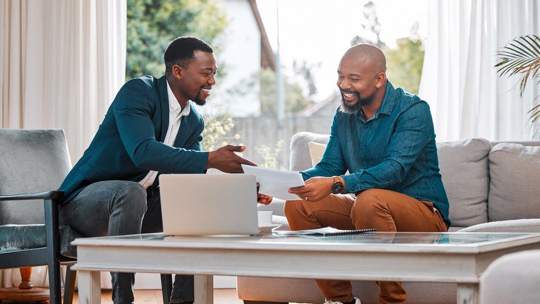 A Chartered Business Valuator sits at a coffee table with his client, pointing to a document while they both smile and look pleased with the discussion.