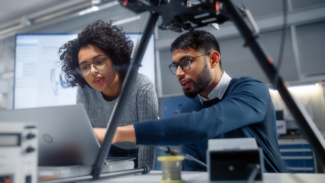 A male engineer points to a laptop while a female engineer looks over his shoulder at what he is gesturing to. In the foreground and background, the technology of their start-up business is on display.