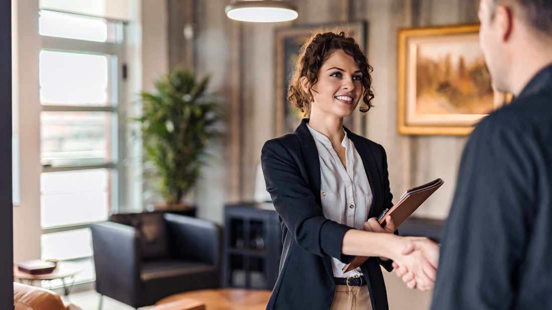 A female real estate agent shakes her client's hand in the living room of a modern house.