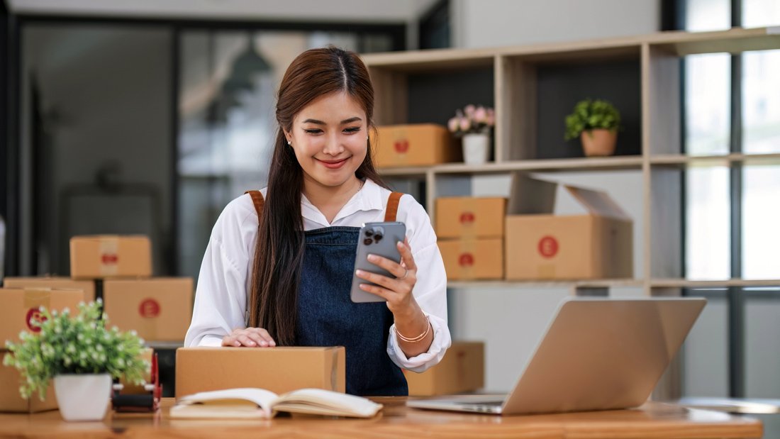 A smiling business owner packaging products while using a smartphone and laptop, representing online sales and digital platform operators.
