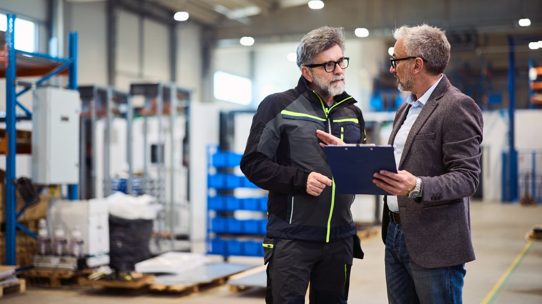 Two professionals discussing business operations on the floor of a manufacturing facility, one holding a clipboard.