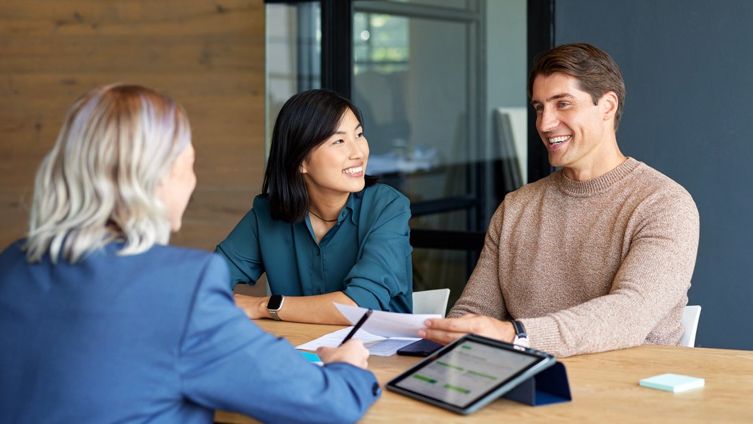 Three people meeting in a modern office to review financial documents, smiling and engaged in discussion.