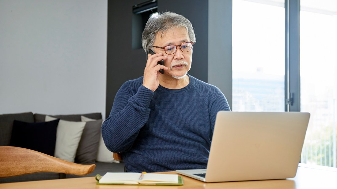 Older man speaking on the phone while looking at a laptop, with a notebook open in front of him at a home office table.