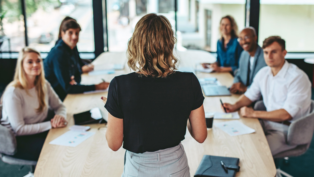 A businesswoman presents to a group of colleagues seated around a conference table during a meeting.