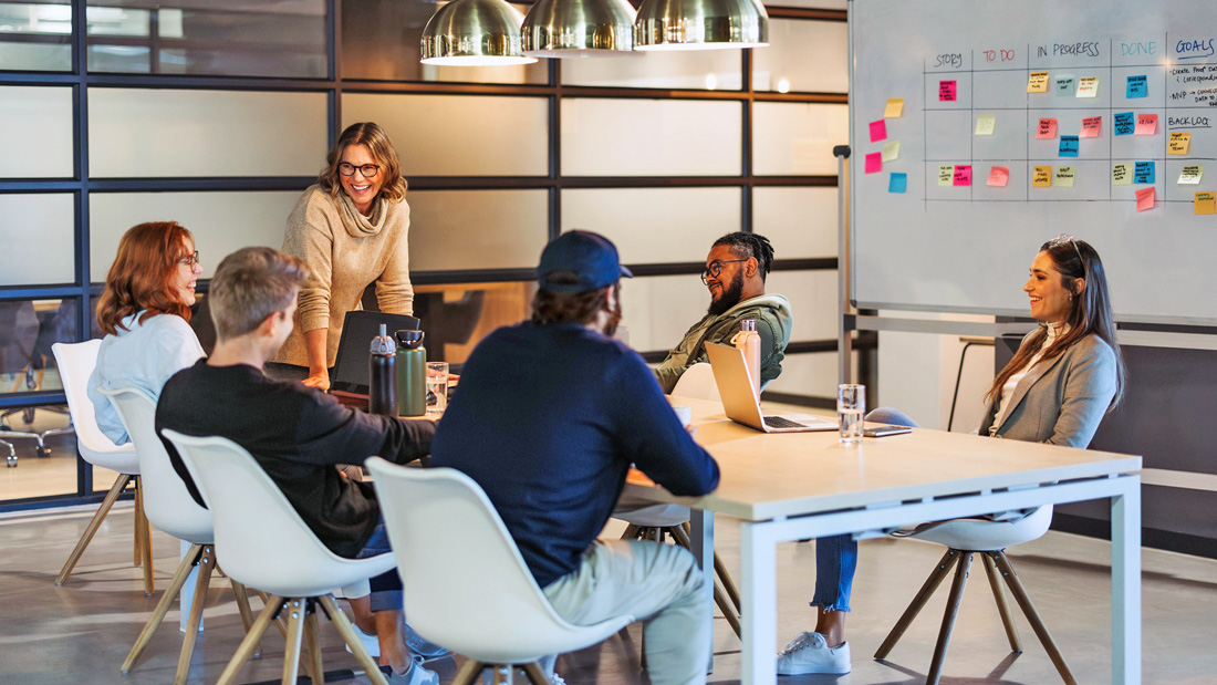 A group of entrepreneurs and advisors meet in a modern office, discussing business growth strategies around a table with laptops and sticky notes on a whiteboard.