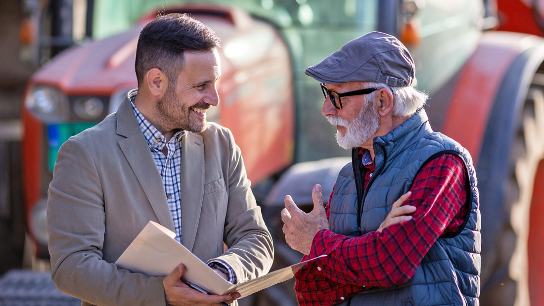 A business advisor speaking with an older client on a farm, with agricultural equipment in the background.